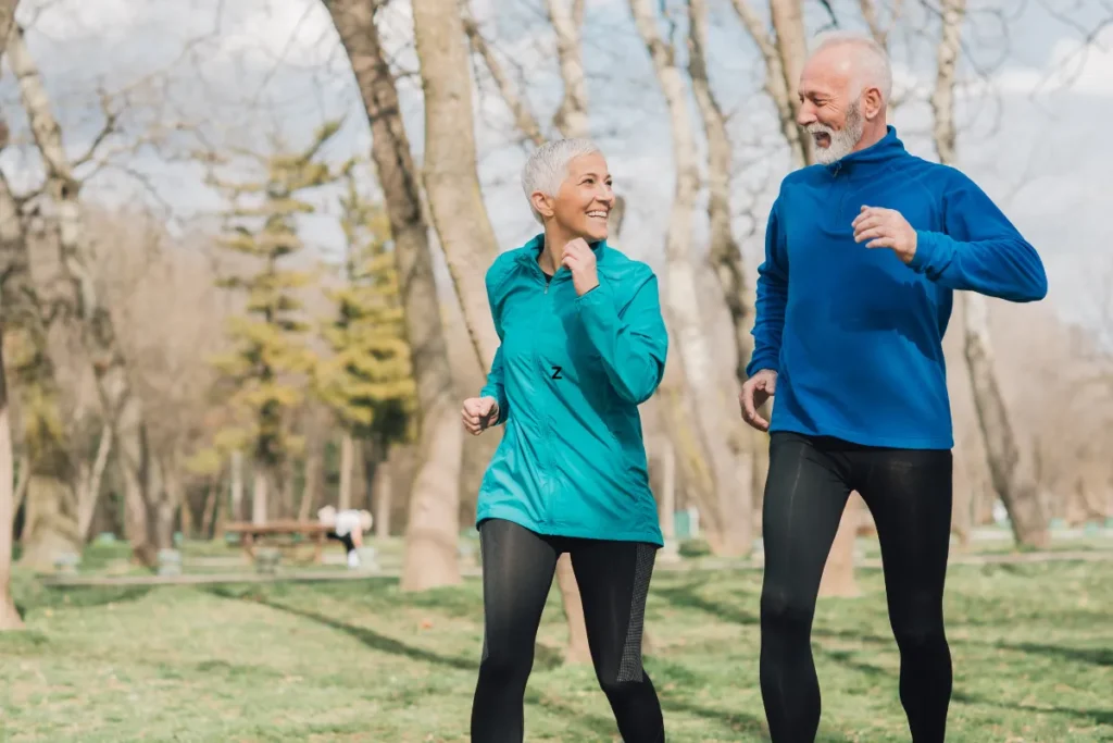 Aktives Seniorenpaar beim Joggen im Frühlingspark, sie in türkiser und er in blauer Sportjacke, beide lächelnd zwischen Birken
