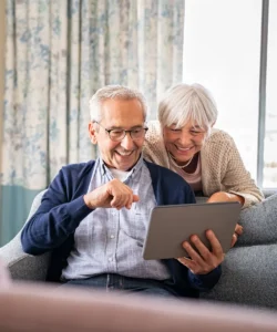 Älteres Paar liest digitalen Newsletter, Mann mit Brille hält Tablet, Frau in beiger Weste schaut interessiert mit