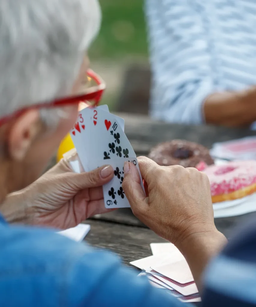 Geistige Fitness durch Kartenspielen, Nahaufnahme von Händen beim Kartenspiel, Spielkarten mit Herz und Kreuz werden gehalten, rosa Donuts im Hintergrund