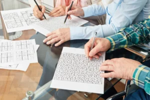 Seniorin beim Gedächtnistraining – geistige Fitness im Alter Gedächtnistraining in der Gruppe. Mehrere Hände beim gemeinsamen Lösen von Denksportaufgaben fürs Gedächtnis, Sudoku und Labyrinthe auf einem Glastisch verteilt.