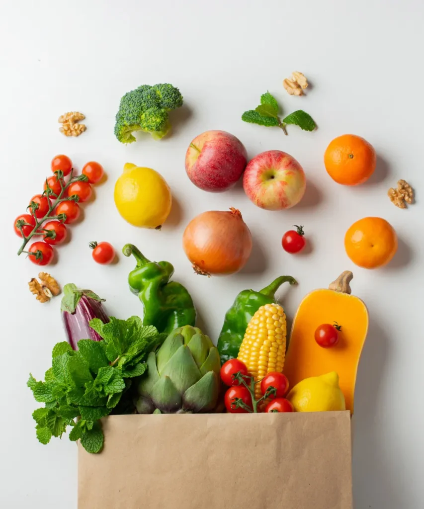 Frische Obst- und Gemüsevielfalt in und um Papiertüte: Tomaten, Äpfel, Orangen, Mais, Paprika, Brokkoli, Walnüsse, Minze und Artischocken auf weißem Hintergrund