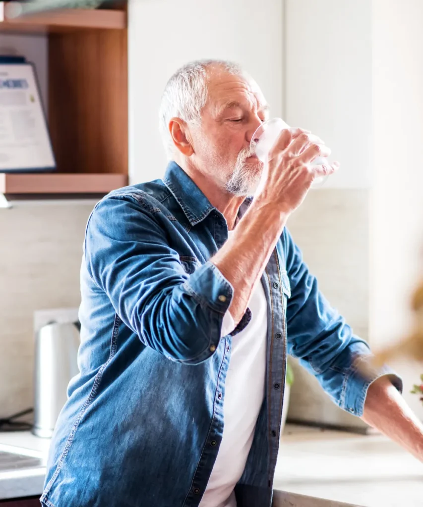 Senior mit grauen Haaren und Jeanshemd trinkt ein Glas Wasser in einer modernen Küche am hellen Tag