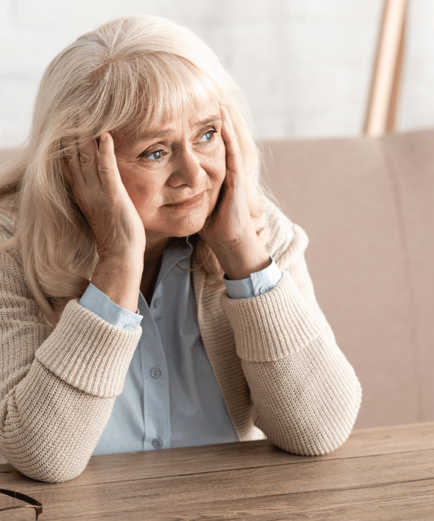 Seniorin mit langen graublonden Haaren sitzt mit den Händen am Kopf an einem Holztisch, trägt hellblaue Bluse und beigene Strickjacke, im Hintergrund ein beiges Sofa