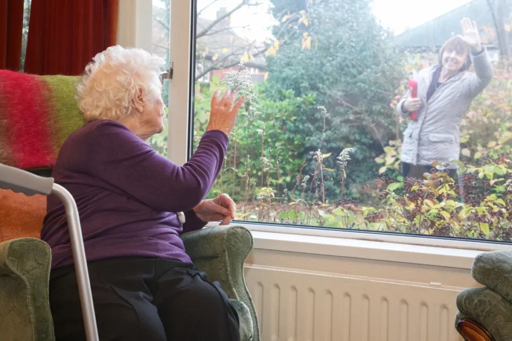 Seniorin mit weißem Haar sitzt im Sessel am Fenster und winkt einer Person im Garten, die draußen zurückwinkt