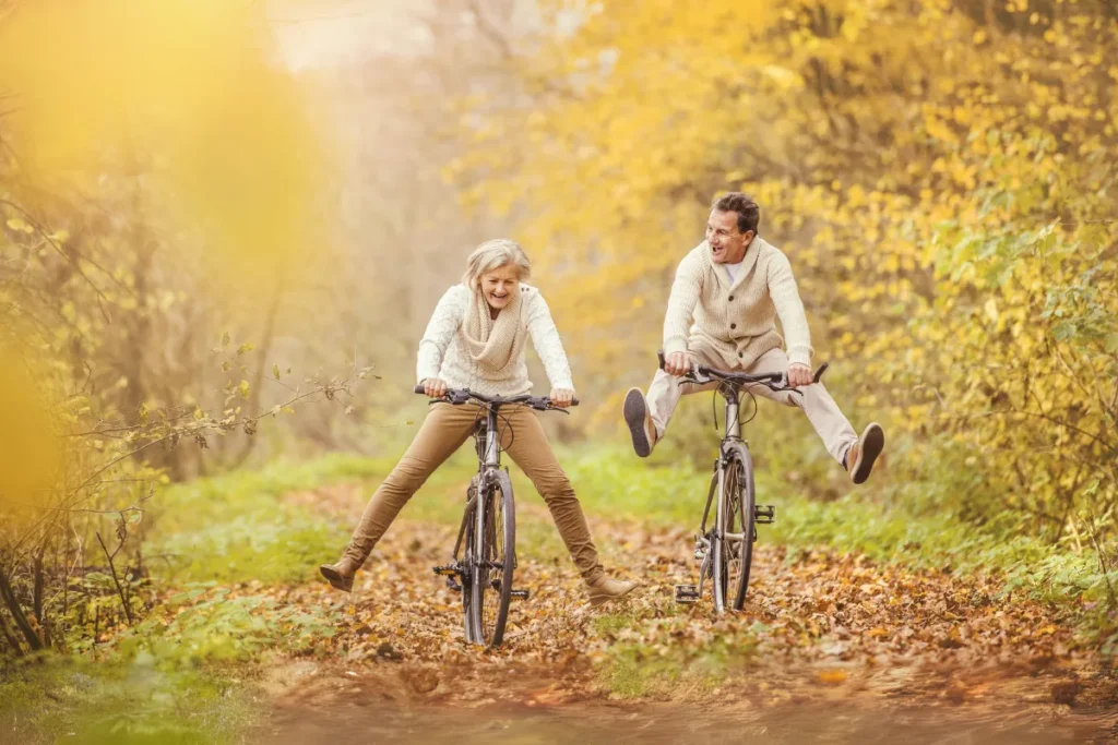 Sportarten für Gehirngesundheit: Zwei Personen in beigen Strickpullovern fahren lachend mit dem Fahrrad durch einen herbstlichen Waldweg, beide mit ausgestreckten Beinen und sichtbarem Spaß an der Bewegung