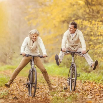 Zwei Personen in beigen Strickpullovern fahren lachend mit dem Fahrrad durch einen herbstlichen Waldweg, beide mit ausgestreckten Beinen und sichtbarem Spaß an der Bewegung