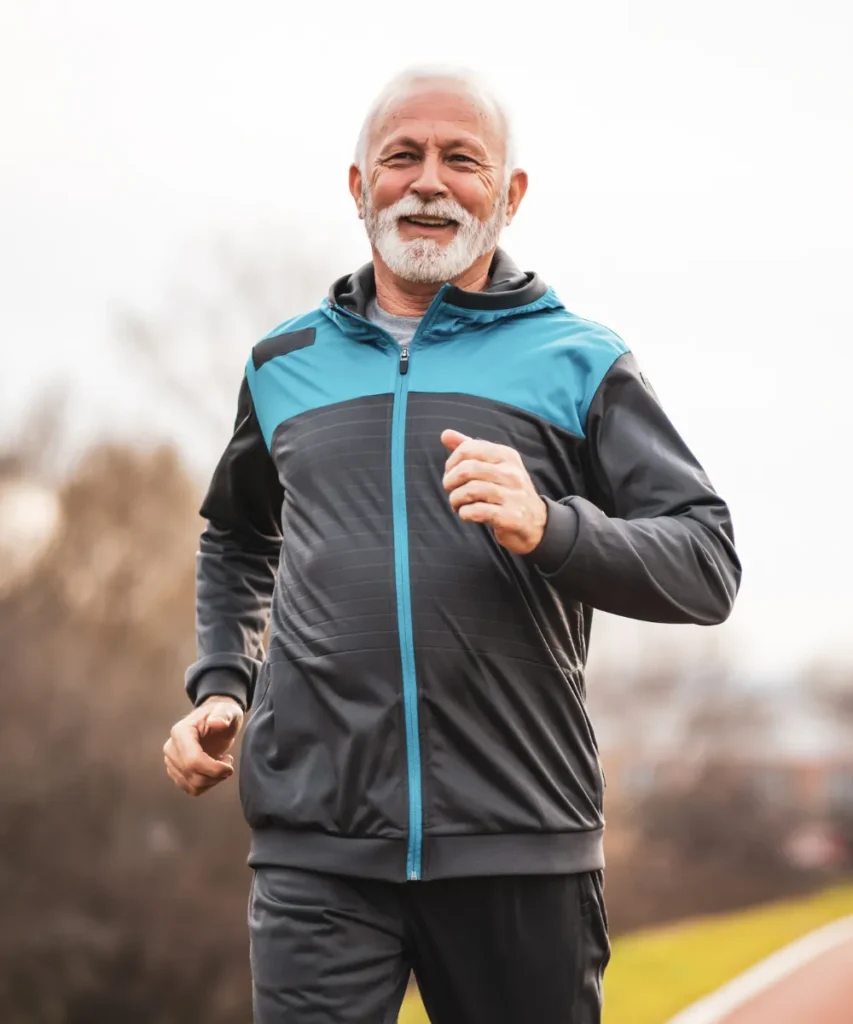 :Sportarten für Gehirngesundheit im Alter: Senior in dunkelgrauer und türkisfarbener Sportjacke joggt auf einer Laufbahn, verschwommene Bäume im Hintergrund