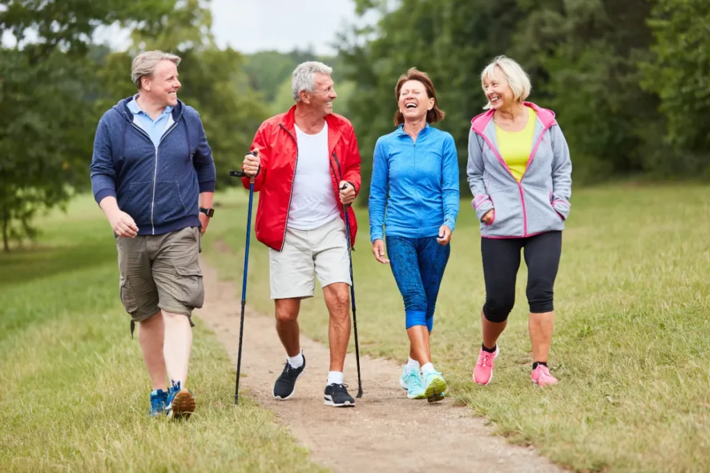 Vier aktive Senioren in sportlicher Kleidung gehen gemeinsam auf einem Naturweg durch eine grüne Parklandschaft, einer mit Nordic-Walking-Stöcken