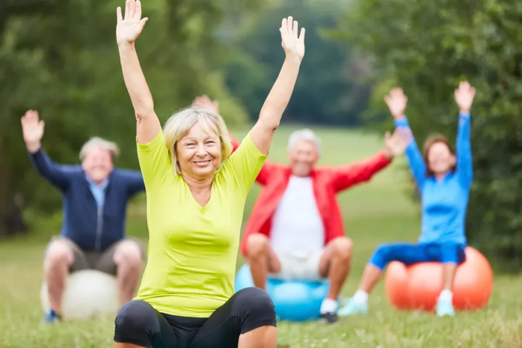 Seniorengruppe macht gemeinsam Gymnastikübungen auf bunten Gymnastikbällen im Park, alle heben die Arme nach oben, grüne Natur im Hintergrund