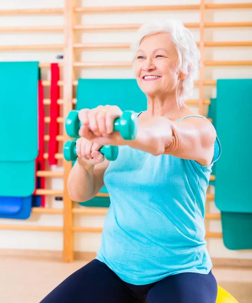 Seniorin in türkisfarbenem Top trainiert mit kleinen Hanteln im Fitnessraum, bunte Matten und Sprossenwand im Hintergrund