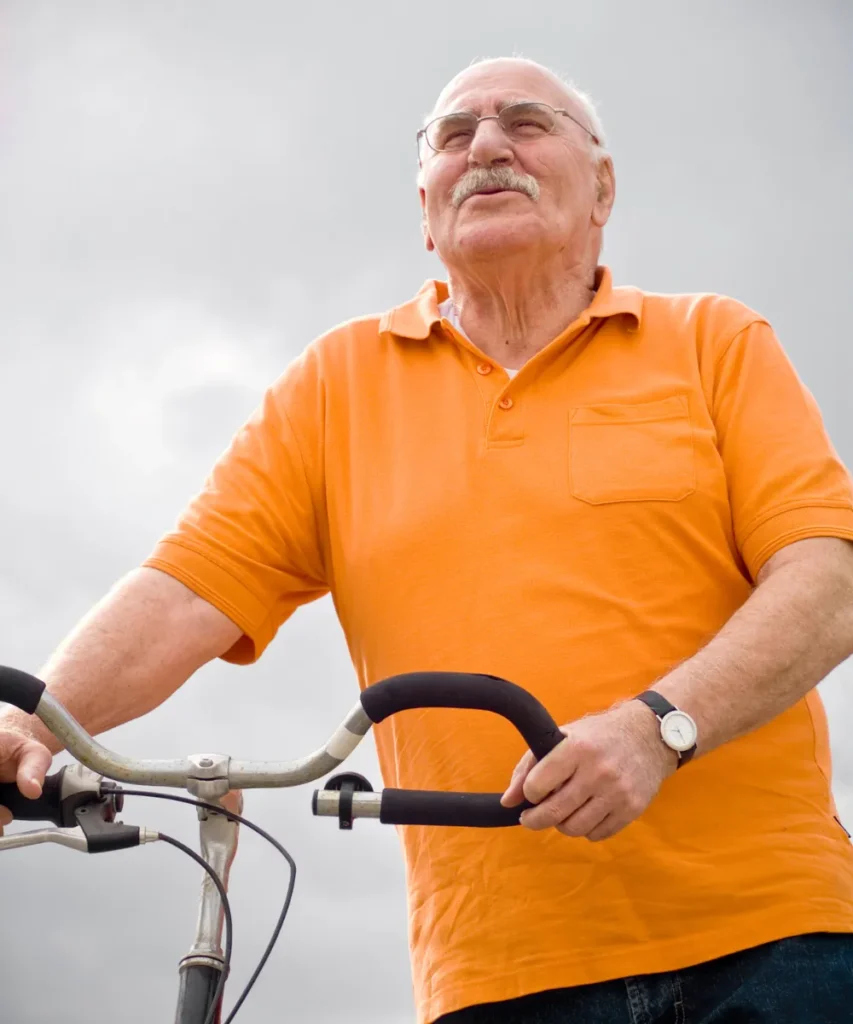 Senior im orangefarbenen Poloshirt hält ein Fahrradlenker fest, vor bewölktem Himmel, Symbol für Mobilität und Bewegung im Alter