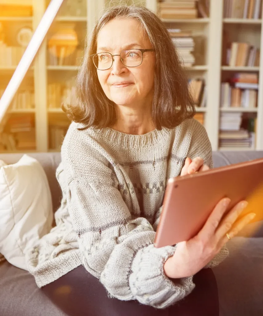 Seniorin mit Brille nutzt entspannt ein Tablet auf dem Sofa vor Bücherregal, Symbol für digitales Lernen im Alter