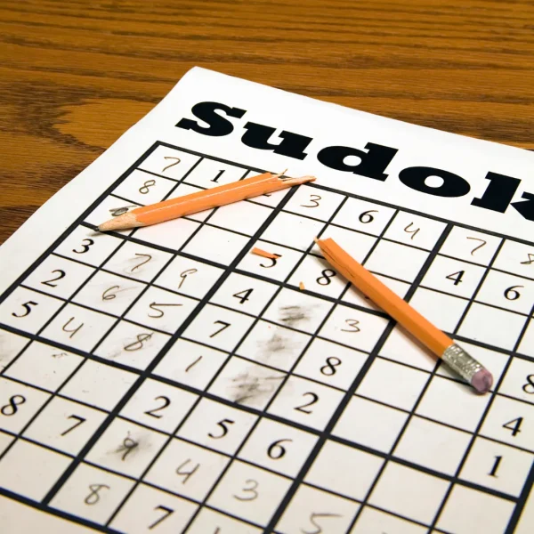 Teilweise ausgefülltes Sudoku-Rätsel auf weißem Papier mit zwei orangefarbenen Bleistiften auf Holzuntergrund